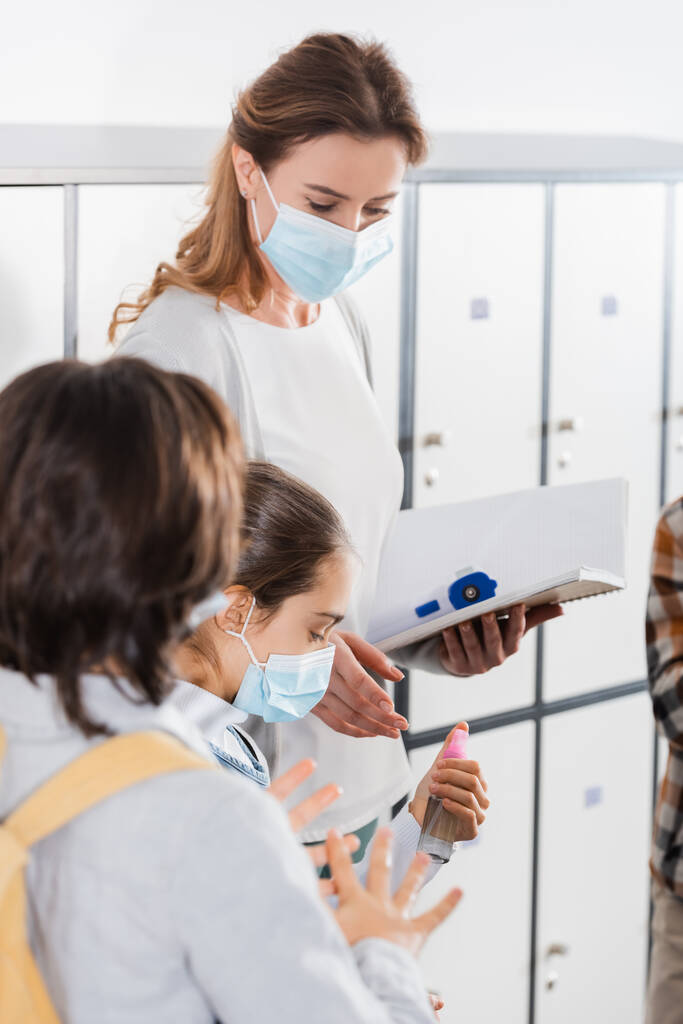 Teacher with notebook and infrared thermometer standing near pupils in protective masks with disinfector  - Photo, Image