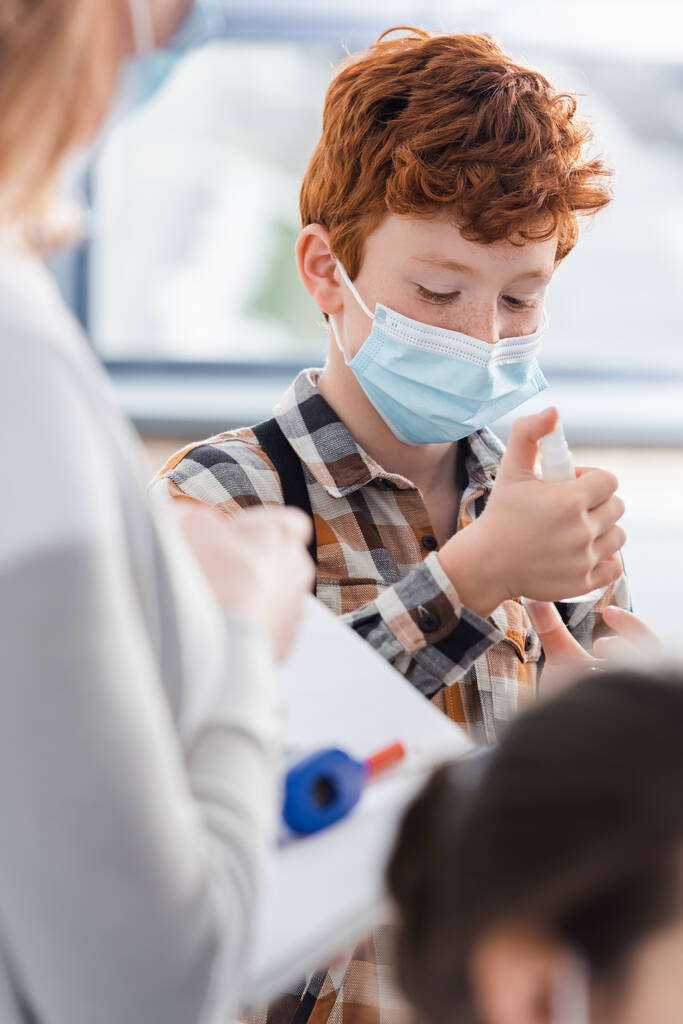 Schoolboy in protective mask using hand sanitizer near teacher on blurred foreground  - Photo, Image
