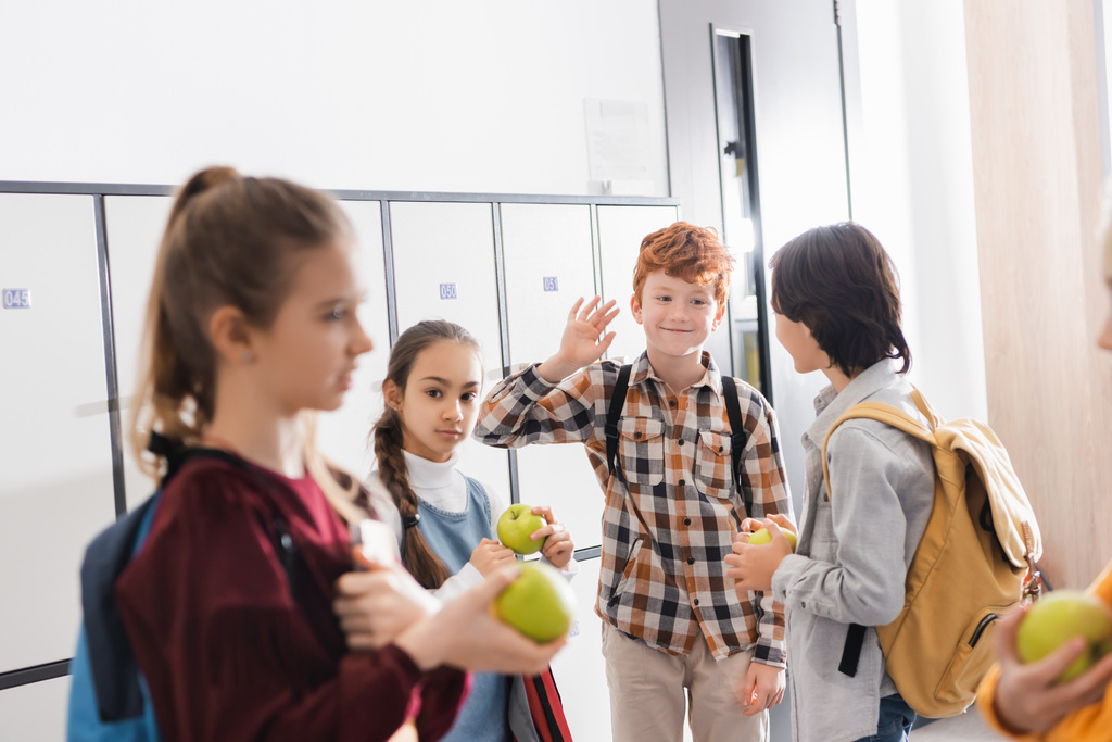 Écolier souriant agitant la main près des amis avec des pommes sur le premier plan flou dans la salle de l'école  - Photo, image