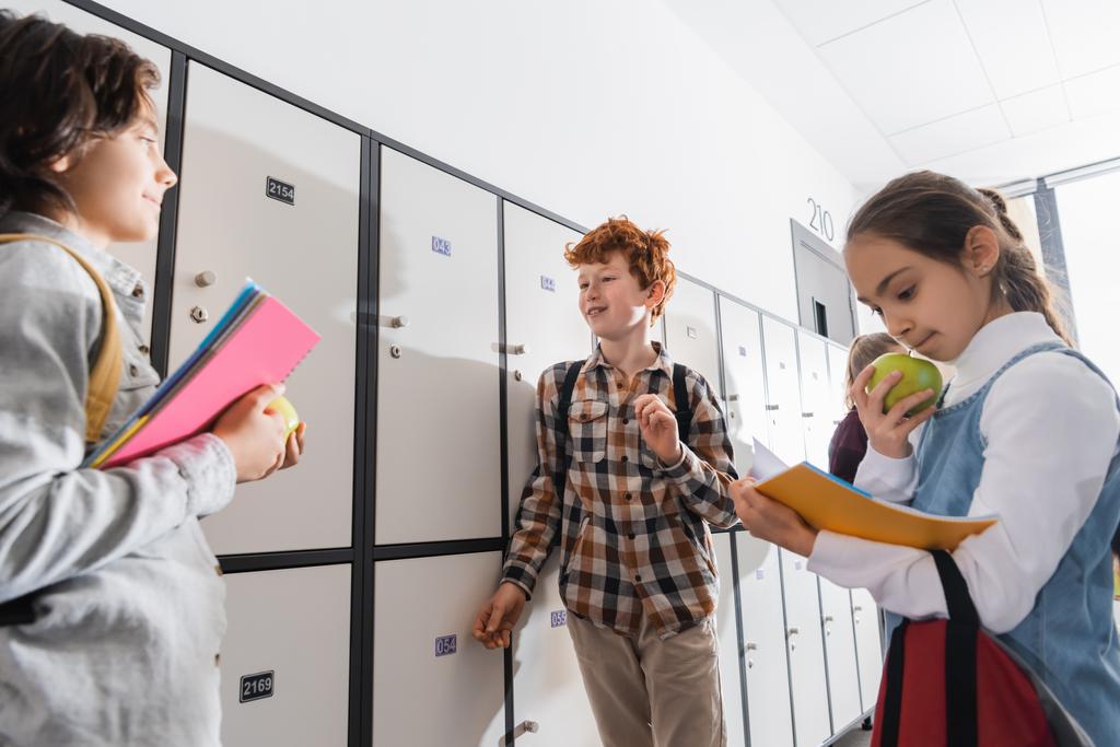 Cheerful boy with backpack standing near lockers and friends with apples and notebooks on blurred foreground in school  - Photo, Image