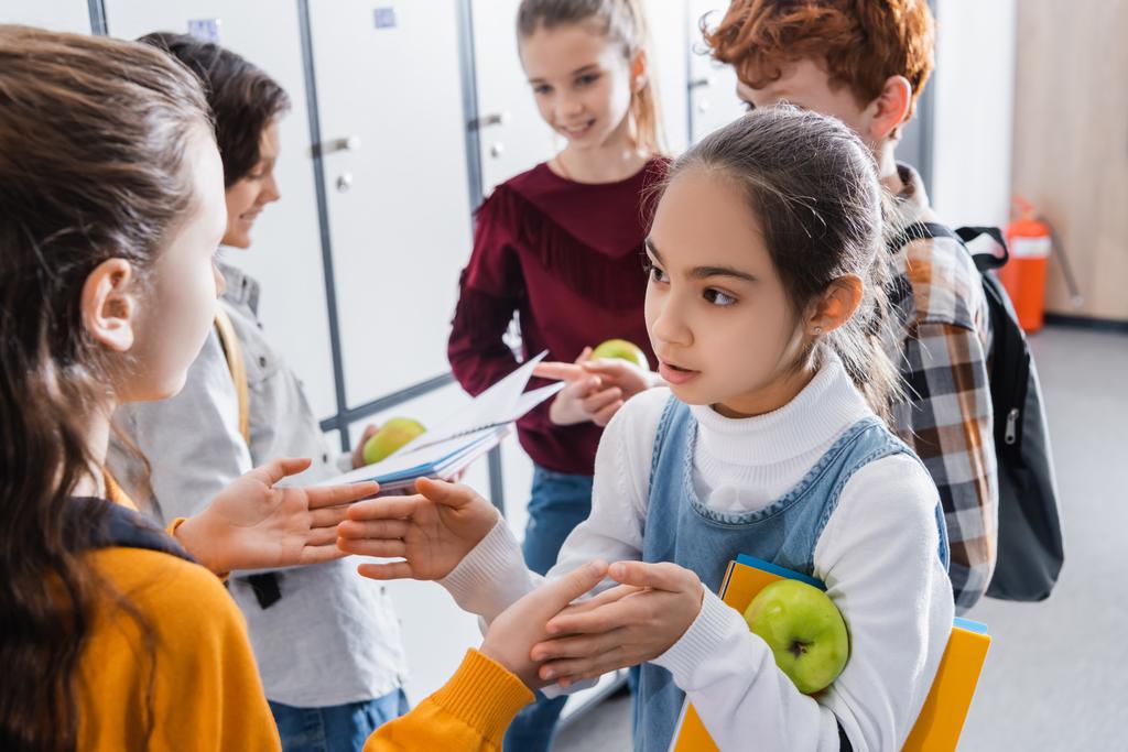 Девочки играют в игры с пирожными рядом с одноклассниками с яблоками и блокнотами на размытом фоне в школе  - Фото, изображение