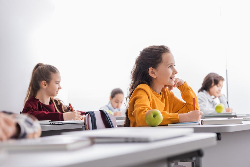 Позитивный ребенок пишет на копировальной книге около спелого яблока и одноклассники на размытом переднем плане  - Фото, изображение