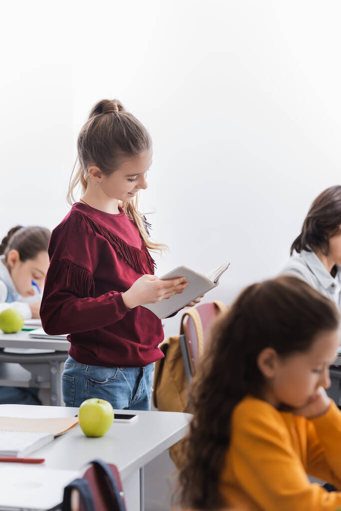 Fröhliche Schülerin liest Buch neben Klassenkameraden im Klassenzimmer  - Foto, Bild