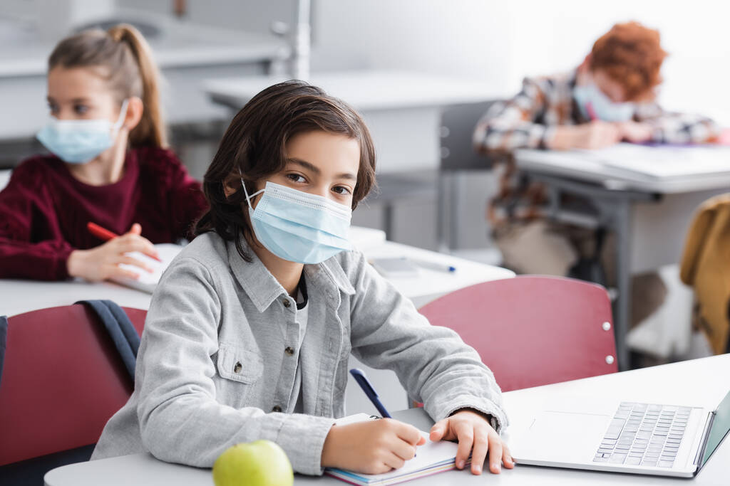 Pupil in medical mask looking at camera while writing on notebook near laptop in classroom  - Photo, Image