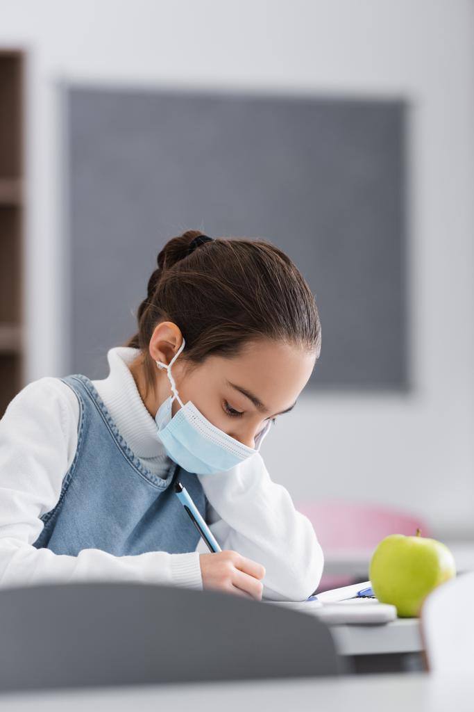 Mädchen mit medizinischer Maske schreibt auf Notizbuch neben Apfel auf Schreibtisch in der Schule  - Foto, Bild