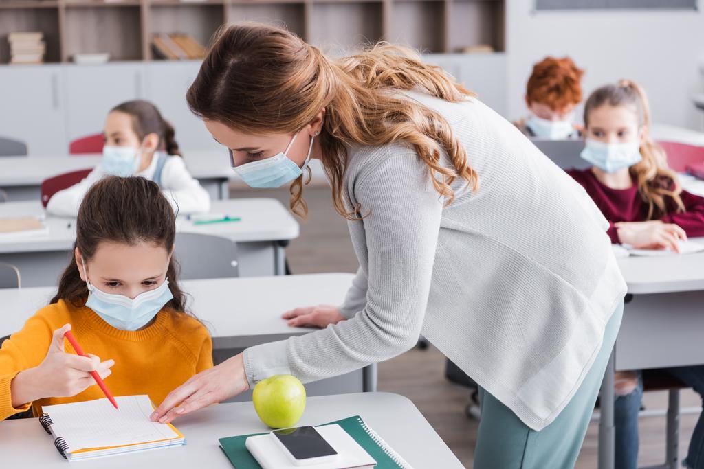 professeur en masque médical regardant portable près de l'écolière pendant la leçon - Photo, image