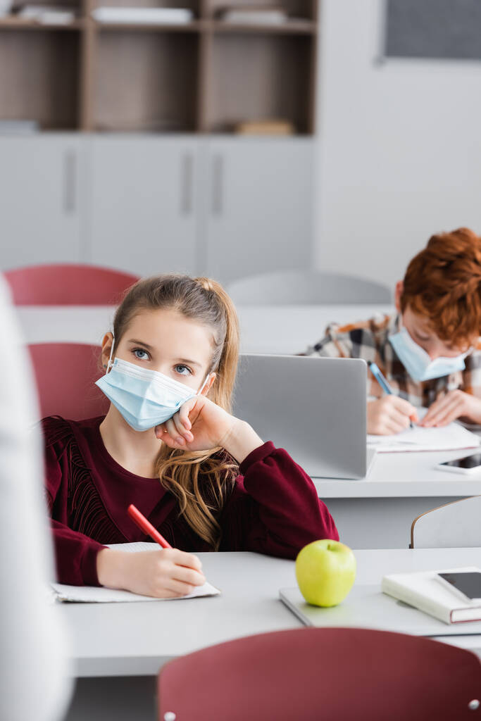 schoolgirl in medical mask holding pen near apple and classmate on blurred background - Photo, Image