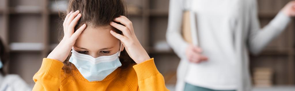 tired schoolgirl in medical mask touching head near teacher on blurred background, banner - Photo, Image