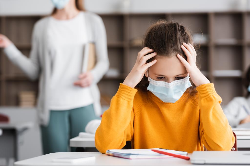 exhausted schoolgirl in medical mask touching head during lesson - Photo, Image