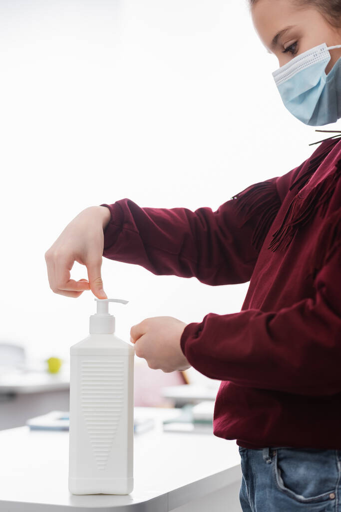 боковой вид школьницы в медицинской маске, наносящей антисептик на руки в классе - Фото, изображение