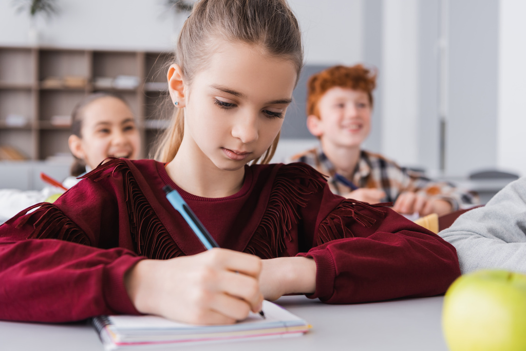 schoolmeisje schrijven in notebook tijdens les op wazig voorgrond - Foto, afbeelding