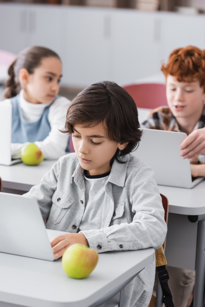 escolar concentrado cerca de la computadora portátil y manzana en el aula - Foto, Imagen