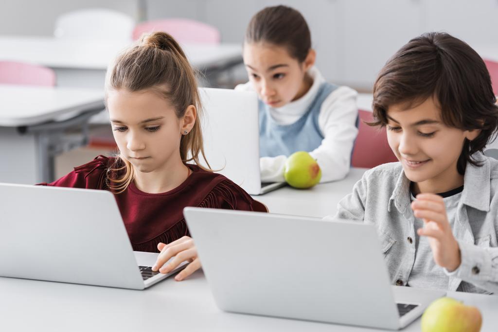 συγκεντρωμένα παιδιά που χρησιμοποιούν φορητό υπολογιστή κατά τη διάρκεια του μαθήματος στο σχολείο σε θολή φόντο - Φωτογραφία, εικόνα
