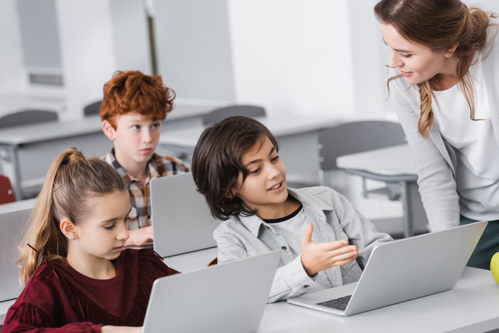 mosolygó iskolás fiú mutatva laptop közelében tanár osztályteremben - Fotó, kép