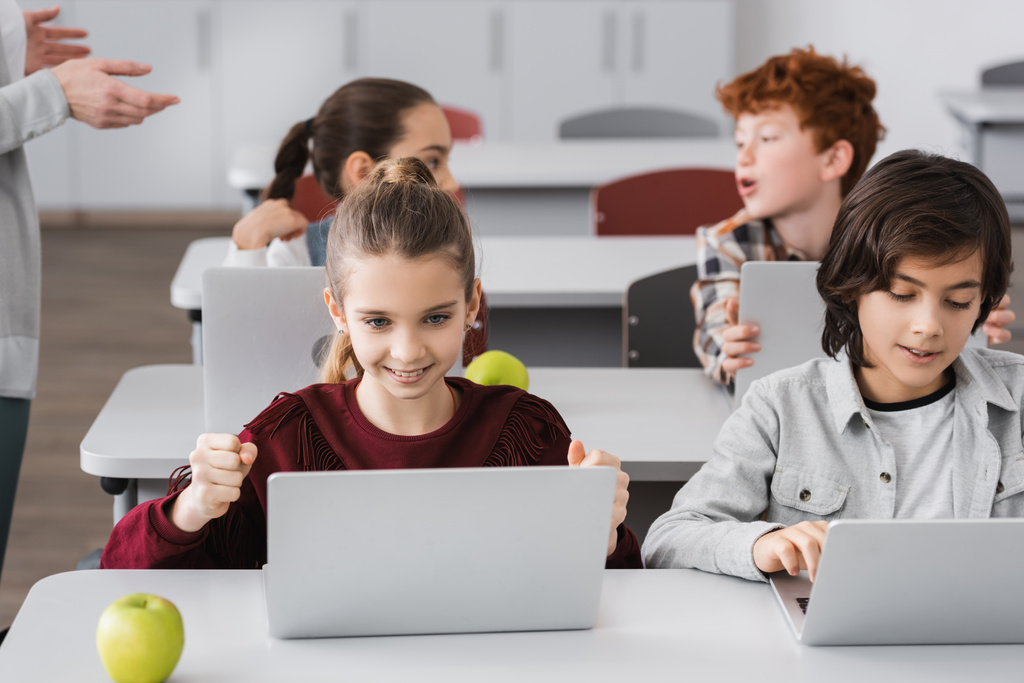 opgewonden meisje tonen win gebaar in de buurt van laptop en klasgenoten op wazig achtergrond - Foto, afbeelding