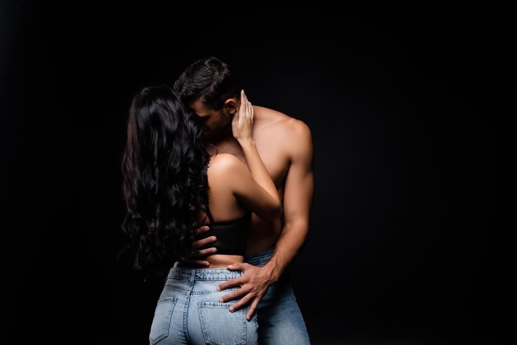 Hombre sin camisa besando morena novia en sujetador y jeans aislados en negro - Foto, Imagen
