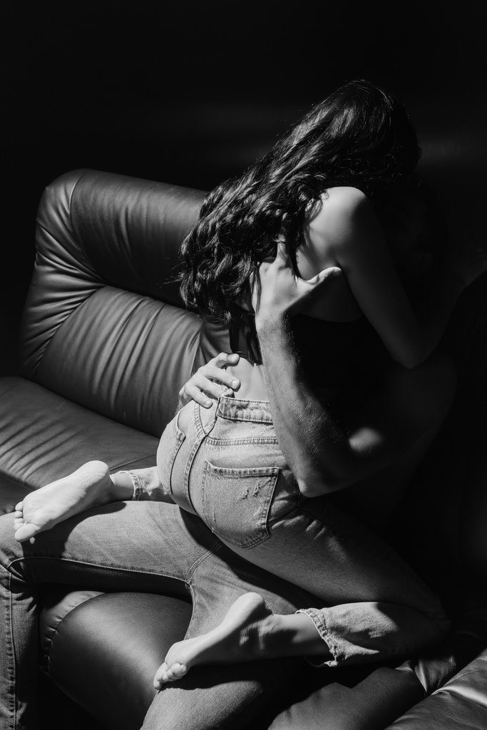 Monochromatyczne ujęcie mężczyzny przytulającego zmysłową kobietę na skórzanej kanapie odizolowaną na czarno  - Zdjęcie, obraz