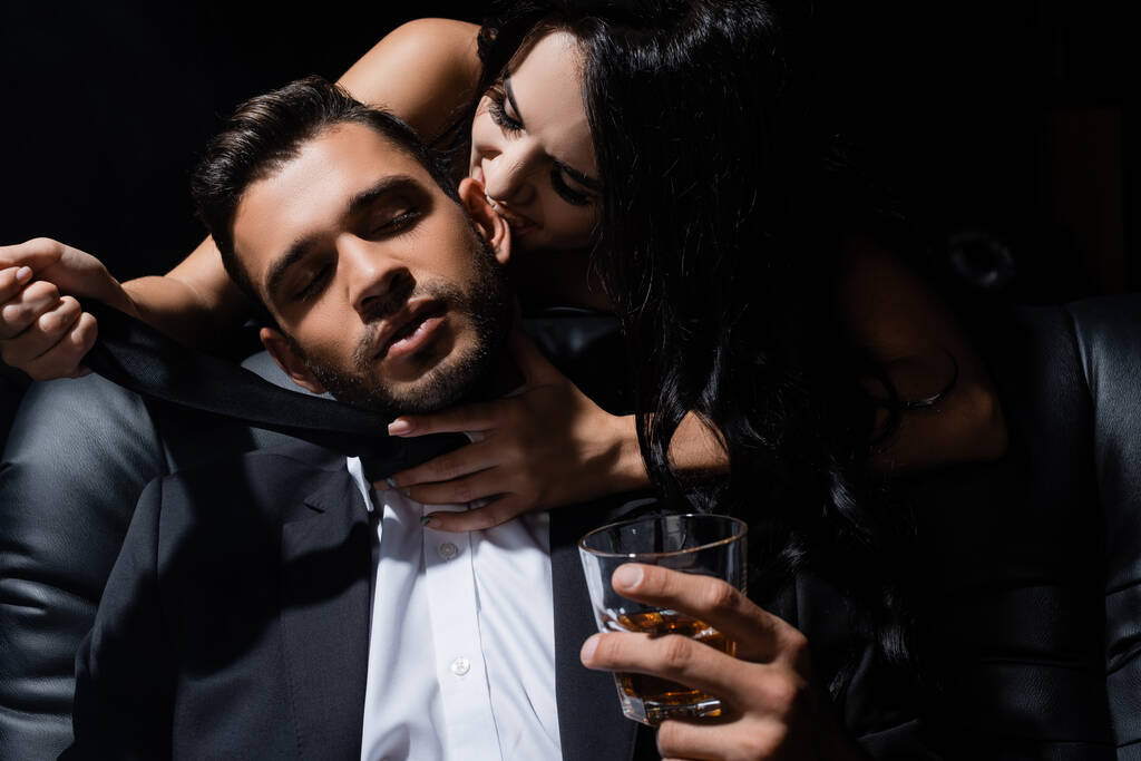 Sexy woman biting ear of boyfriend with whiskey isolated on black  - Photo, Image
