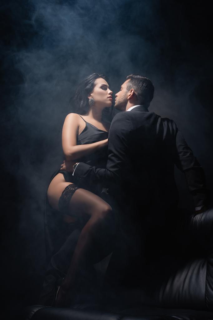 Сексуальна жінка в сукні і панчохах, що сидить біля хлопця в костюмі на дивані на чорному тлі з димом
  - Фото, зображення