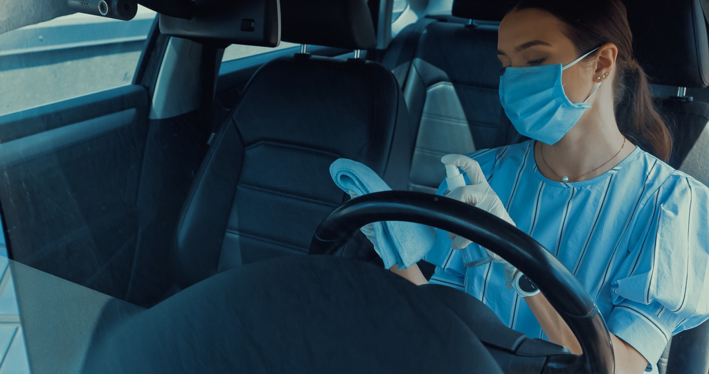 Frau mit medizinischer Maske sprüht Desinfektionsmittel auf Lappen im Auto - Foto, Bild