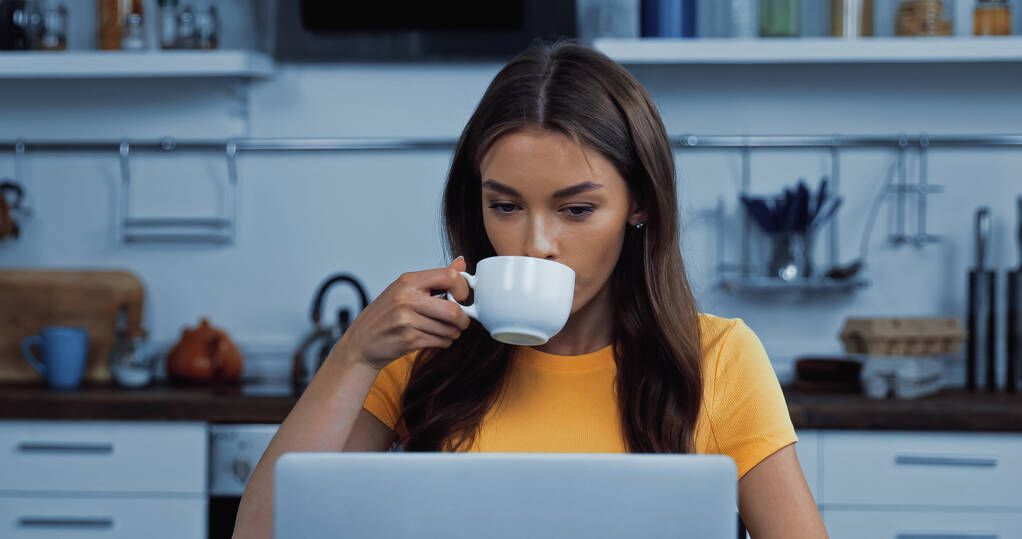 ブルネット若い女性がノートパソコンの近くでコーヒーを飲む  - 写真・画像