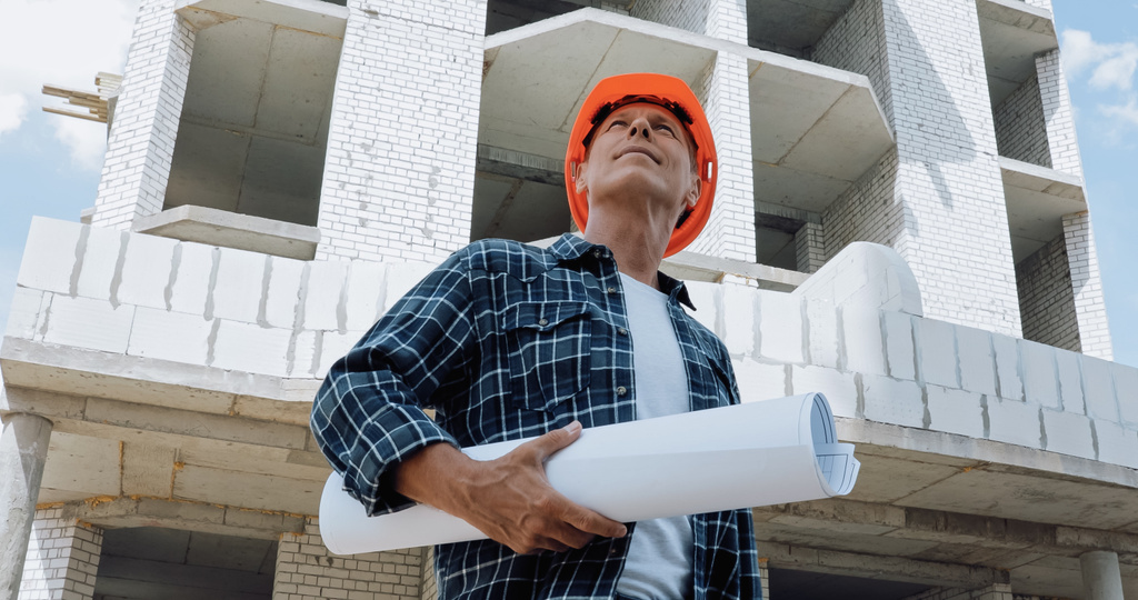 low angle view of mature builder holding blueprint and smiling on construction site - Photo, Image