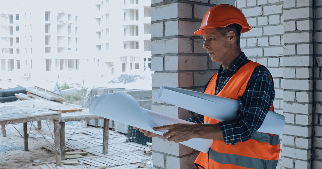 Комплексный строитель в шлеме смотрит на чертеж на строительной площадке - Фото, изображение