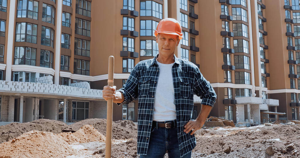 Hymyilevä rakentaja pitelee lapiota ja seisoo käsi lantiolla rakennustyömaalla - Valokuva, kuva