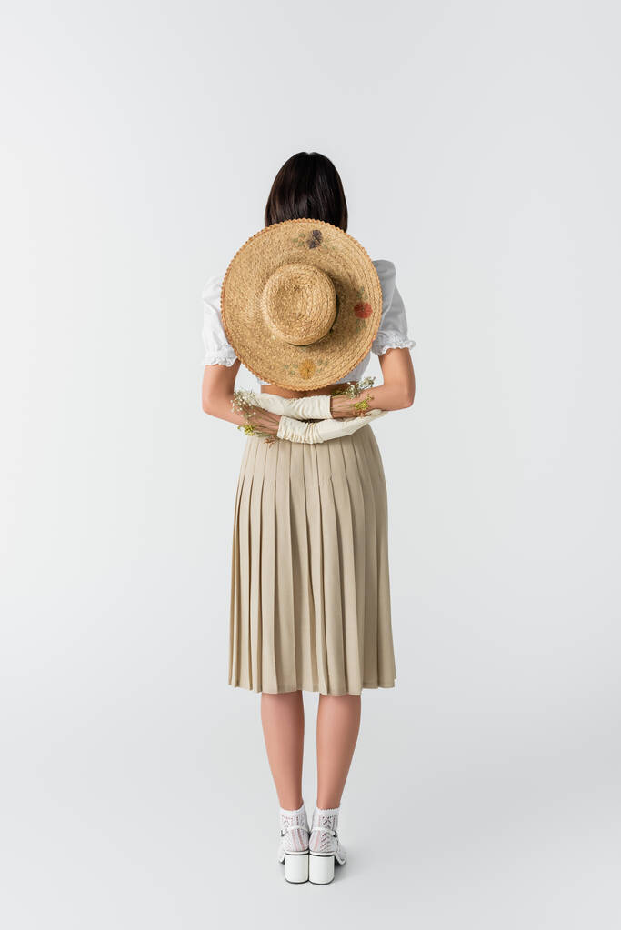 πλήρες μήκος της γυναίκας σε γάντια με λουλούδια και ψάθινο καπέλο πίσω από την πλάτη σε λευκό  - Φωτογραφία, εικόνα