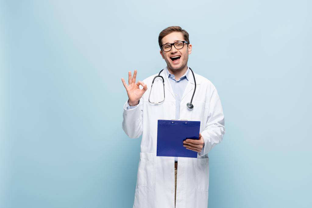 verbaasd jonge arts in bril en witte jas houden klembord en tonen oke teken op blauw - Foto, afbeelding