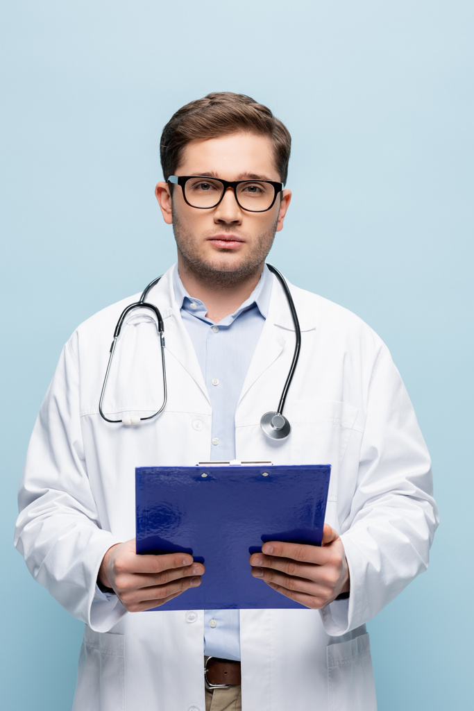 молодой врач в очках и белом халате, держащий планшет и смотрящий на камеру, изолированную на голубом - Фото, изображение