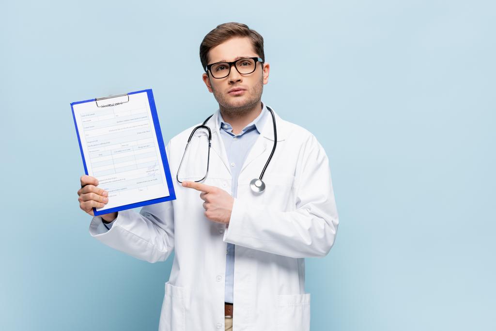 doctor en gafas y abrigo blanco apuntando con el dedo al portapapeles con letras de tarjetas médicas en azul - Foto, Imagen