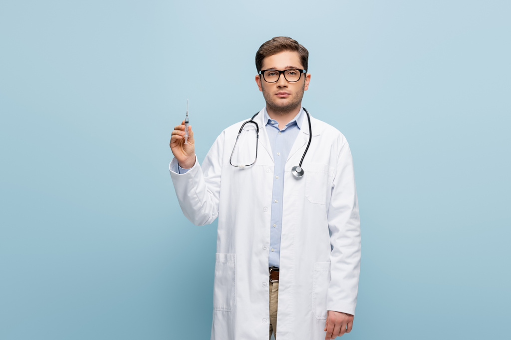 γιατρός σε γυαλιά και λευκό παλτό κρατώντας σύριγγα σε μπλε - Φωτογραφία, εικόνα