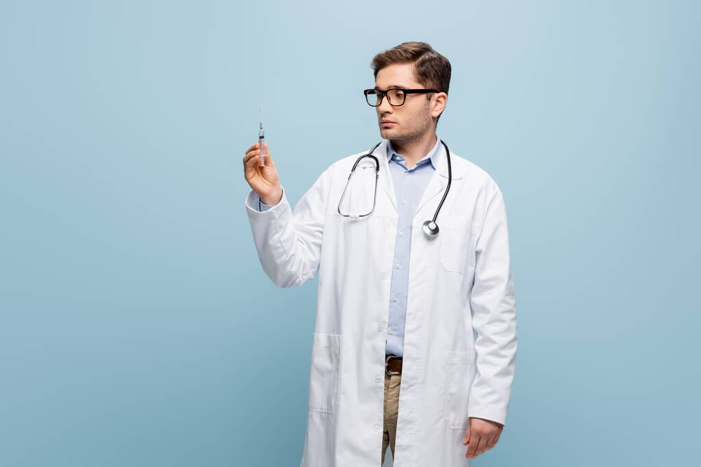 młody lekarz w okularach i biały płaszcz patrzący na strzykawkę na niebiesko - Zdjęcie, obraz