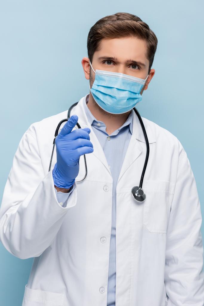 médico en máscara médica y bata blanca con estetoscopio apuntando con el dedo aislado en azul - Foto, Imagen