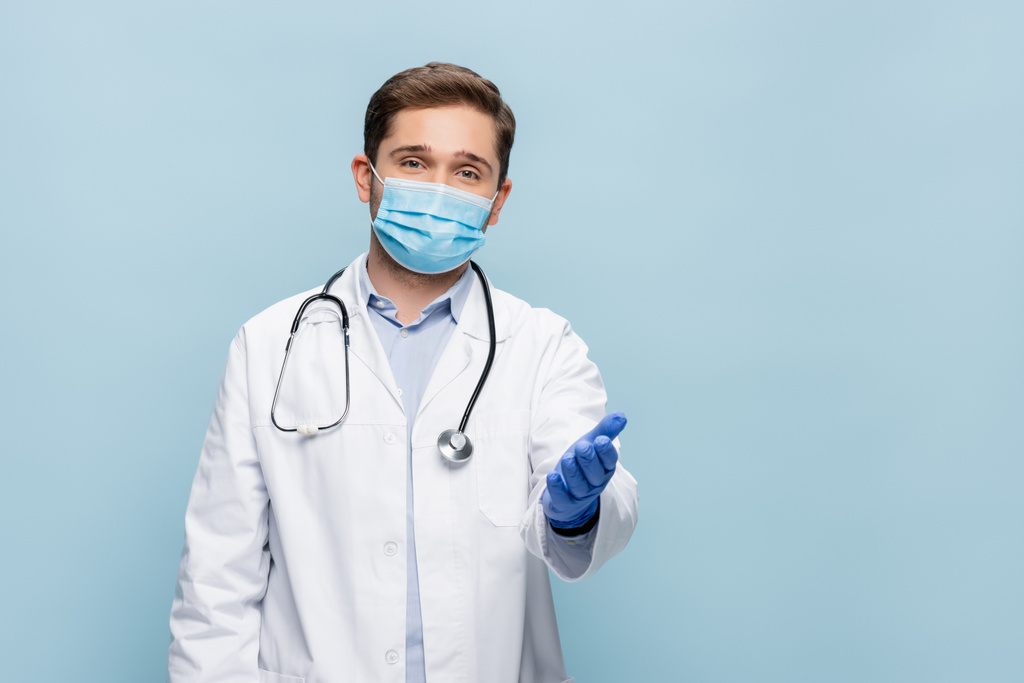 médico en máscara médica y bata blanca con estetoscopio apuntando con la mano aislada en azul - Foto, Imagen
