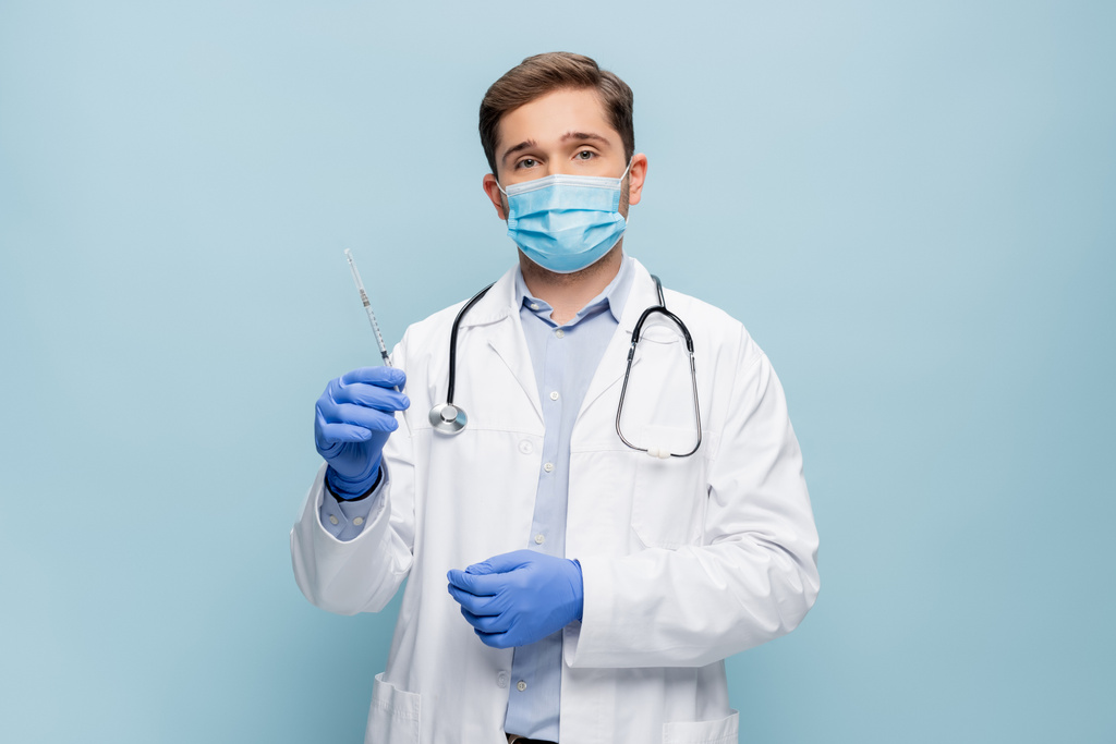 jeune médecin en masque médical et manteau blanc avec stéthoscope tenant la seringue isolée sur bleu - Photo, image