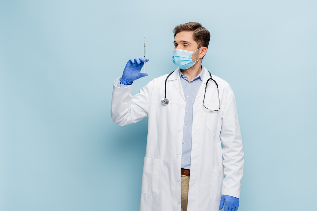 νεαρός γιατρός με ιατρική μάσκα και λευκή επικάλυψη με σύριγγα συγκράτησης στηθοσκοπίου με εμβόλιο στο μπλε - Φωτογραφία, εικόνα