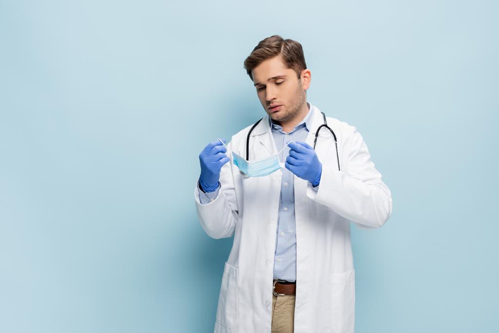 νεαρός γιατρός σε λευκό παλτό με στηθοσκόπιο κρατώντας ιατρική μάσκα σε μπλε - Φωτογραφία, εικόνα
