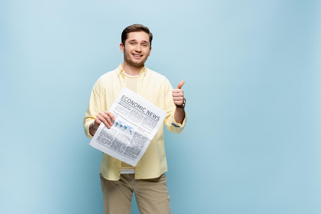 sonriente hombre en camisa sosteniendo periódico y mostrando el pulgar hacia arriba aislado en azul - Foto, Imagen