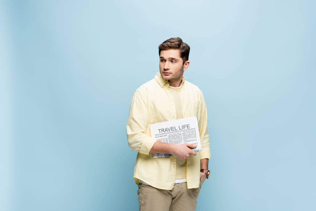 Молодой человек в рубашке держит газету путешествия и смотрит в сторону изолированы на синий - Фото, изображение