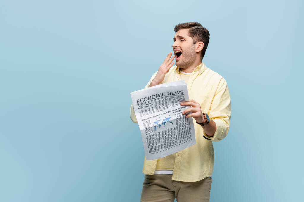 σοκαρισμένος νεαρός άνδρας με ανοιχτό το στόμα κρατώντας εφημερίδα απομονωμένη στο μπλε - Φωτογραφία, εικόνα