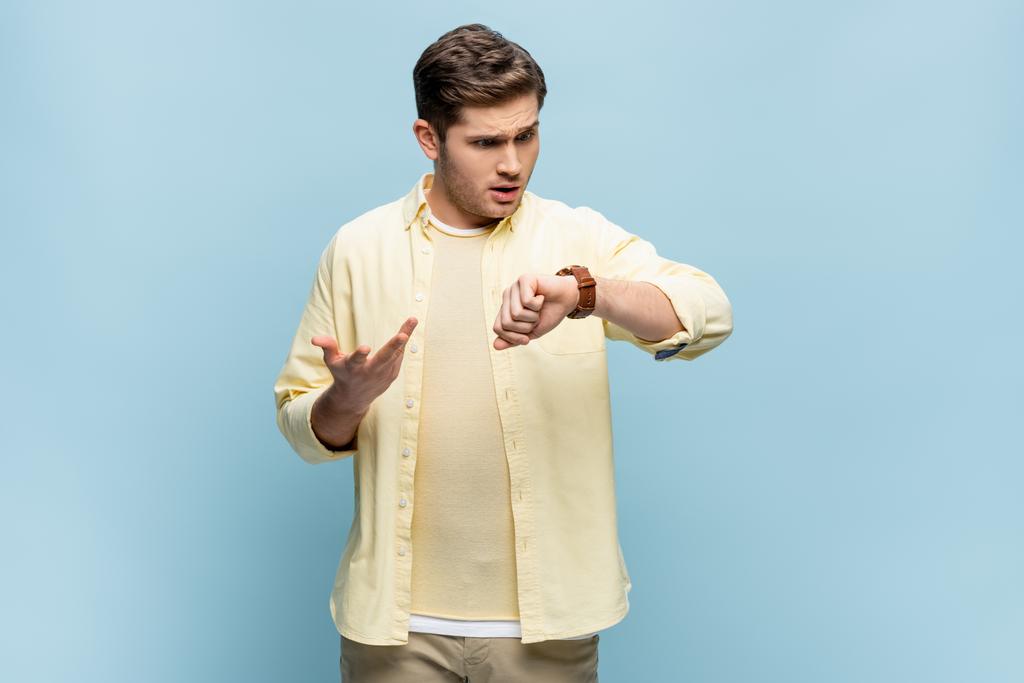 nespokojen mladý muž ve žluté košili při pohledu na hodinky na modré - Fotografie, Obrázek