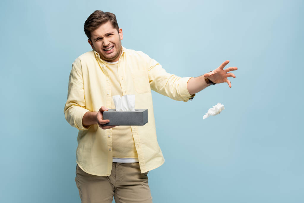 άρρωστος άνθρωπος πετώντας χαρτοπετσέτα μακριά και κρατώντας κουτί ιστού απομονώνονται σε μπλε - Φωτογραφία, εικόνα