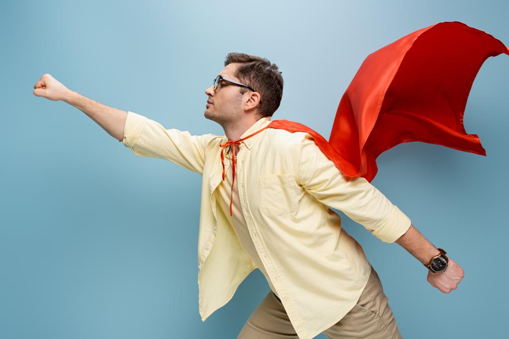 zijaanzicht van superheld in bril en rode cape staande met uitgestrekte hand op blauw - Foto, afbeelding