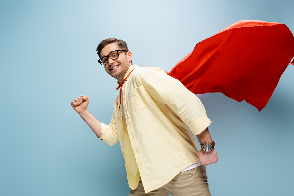 alegre superhéroe en gafas y capa roja de pie con el puño apretado en azul - Foto, Imagen