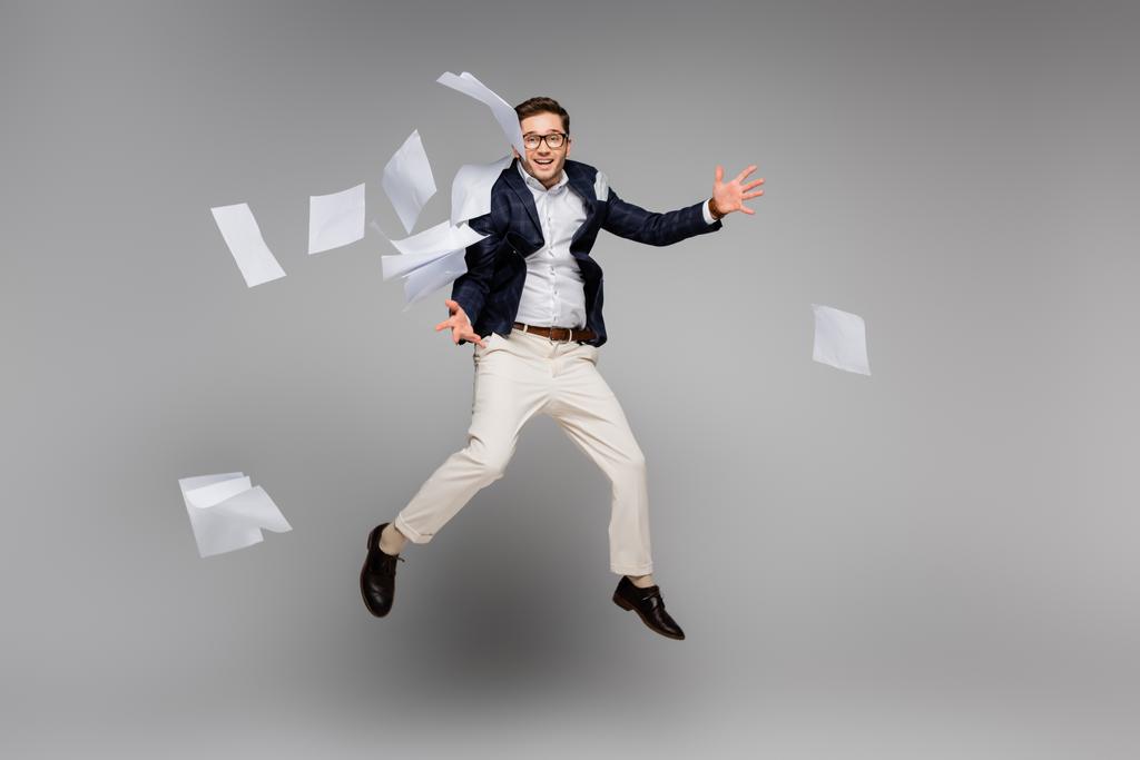 longitud completa de feliz hombre de negocios saltando cerca de papeles en el aire en gris - Foto, Imagen