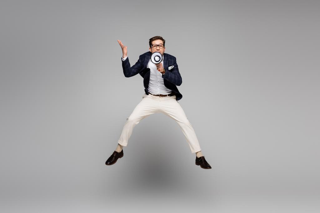 longitud completa de hombre de negocios saltando y hablando en megáfono en gris - Foto, imagen