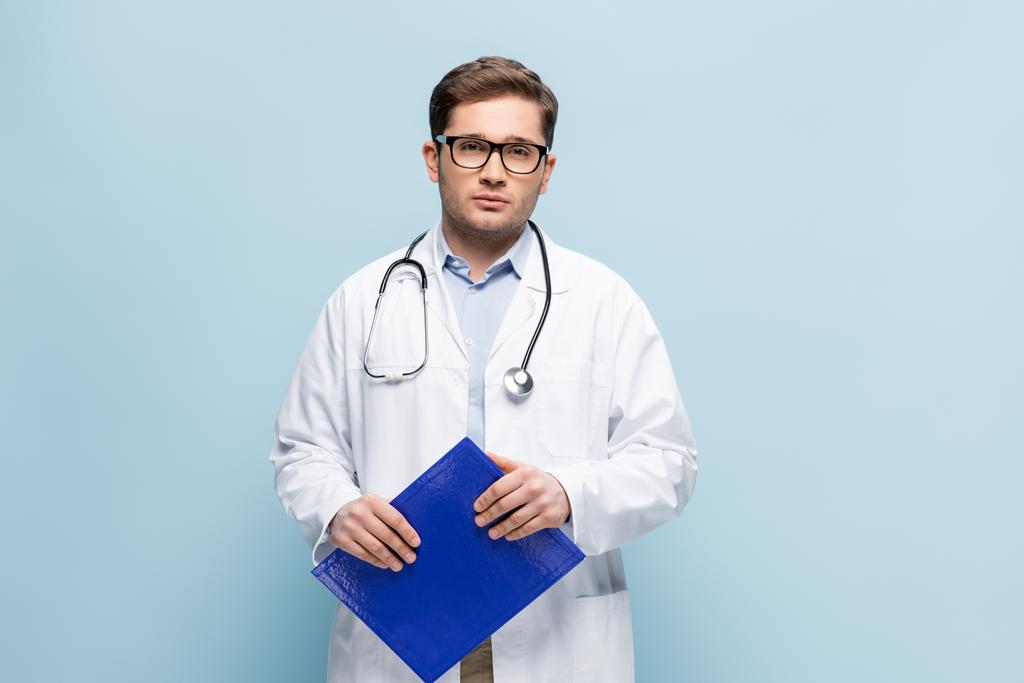 Arzt in weißem Mantel und Brille mit Klemmbrett und Blick auf Kamera isoliert auf blau - Foto, Bild