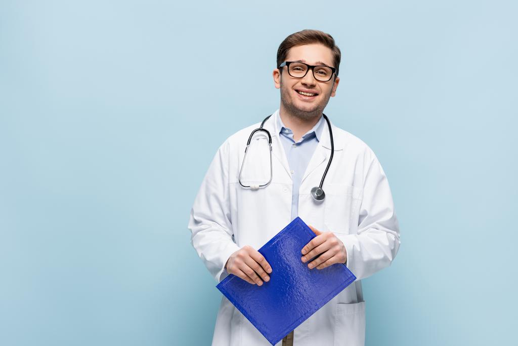 χαμογελαστός γιατρός με γυαλιά και λευκό παλτό κρατώντας πρόχειρο και κοιτάζοντας κάμερα στο μπλε - Φωτογραφία, εικόνα